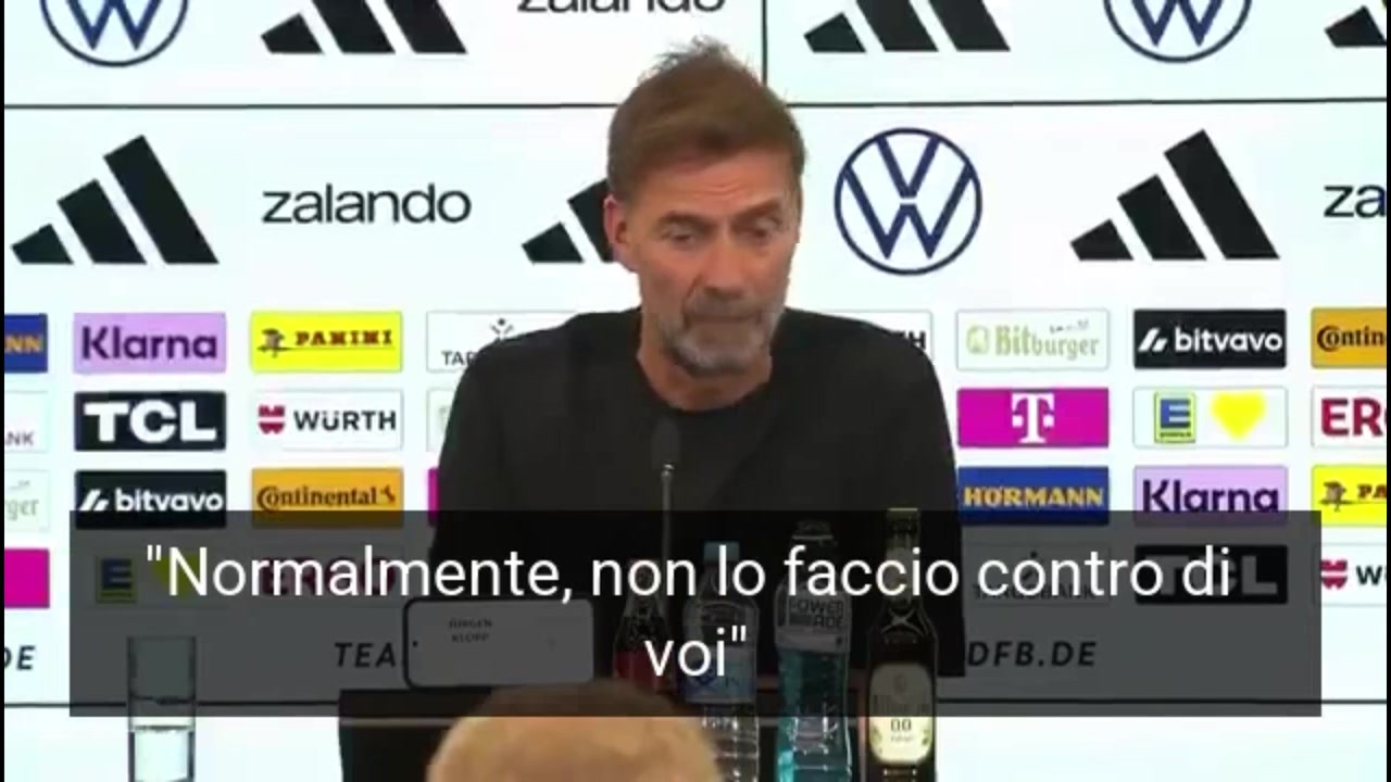 Allenatore tedesco in conferenza stampa con bandiera nazionale sullo sfondo, abito formale, espressione seria, immagine di approfondimento
