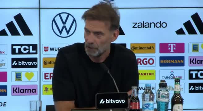 Allenatore tedesco in conferenza stampa con bandiera nazionale sullo sfondo, abito formale, espressione seria