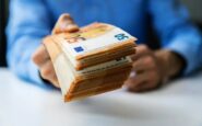Primo piano di una persona con una camicia azzurra che porge in avanti una spessa mazzetta di 50 euro