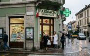 Farmacia comunale con insegna e croce verde luminosa all'angolo di una strada urbana. Davanti all'ingresso, una farmacista in camice bianco assiste un'anziana signora.