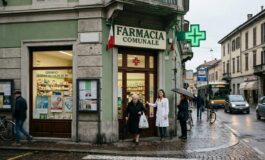 Farmacia comunale con insegna e croce verde luminosa all'angolo di una strada urbana. Davanti all'ingresso, una farmacista in camice bianco assiste un'anziana signora.