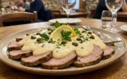 Piatto ovale di vitello tonnato a fette sottili coperto da salsa tonnata cremosa, decorato con capperi, prezzemolo fresco e una fetta di limone, servito su un tavolo di legno