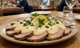 Piatto ovale di vitello tonnato a fette sottili coperto da salsa tonnata cremosa, decorato con capperi, prezzemolo fresco e una fetta di limone, servito su un tavolo di legno