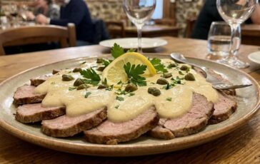 Piatto ovale di vitello tonnato a fette sottili coperto da salsa tonnata cremosa, decorato con capperi, prezzemolo fresco e una fetta di limone, servito su un tavolo di legno