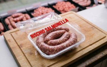 Vassoio di plastica con salsiccia fresca a spirale su tagliere di legno in macelleria, con scritta in sovrimpressione rischio salmonella